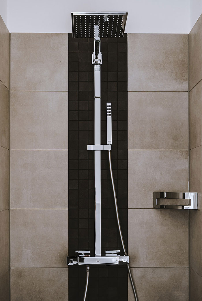 Moderne Dusche mit Regenduschkopf, Handbrause und großen beigen Fliesen sowie dunklem Mosaikstreifen an der Wand