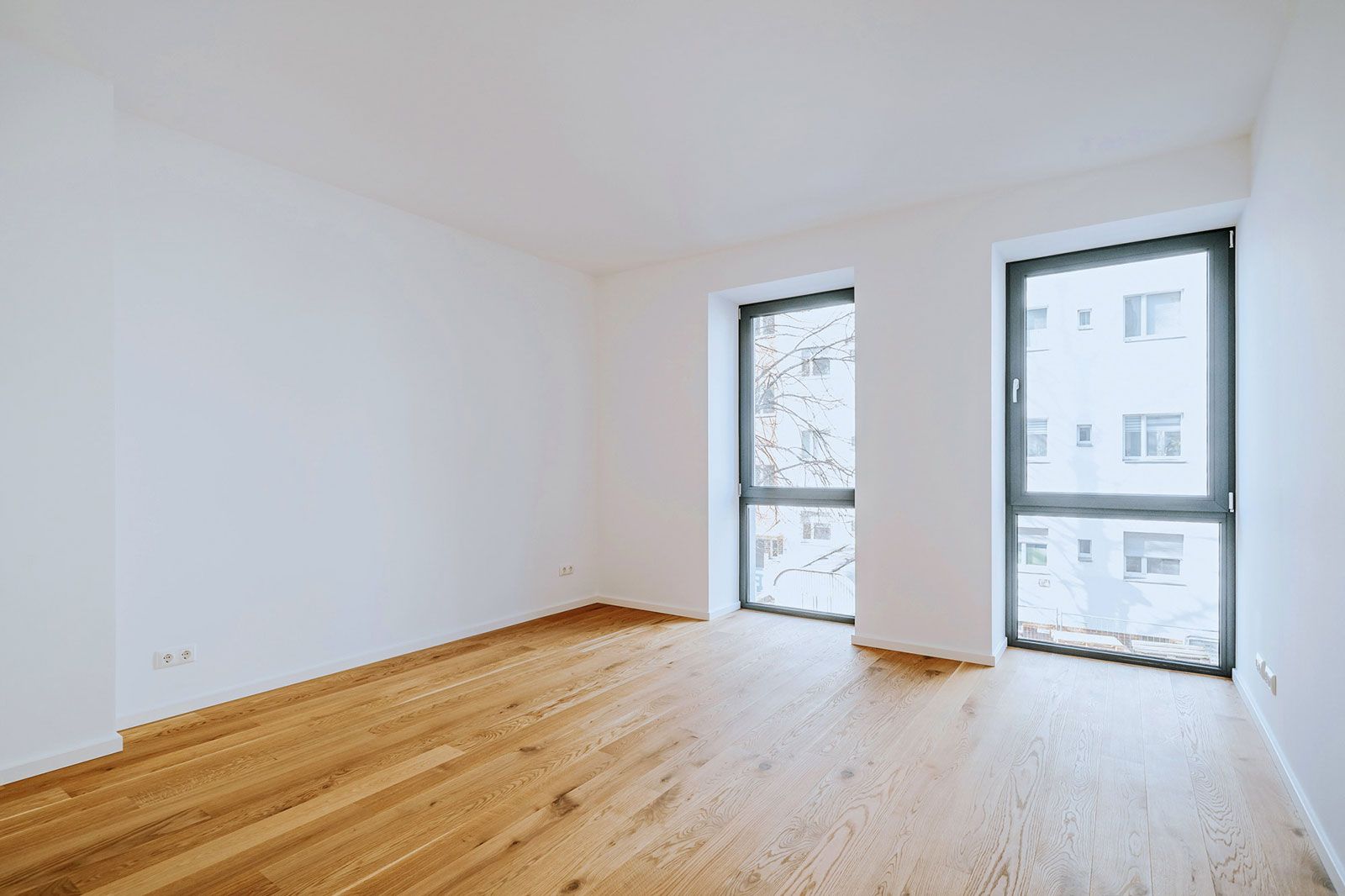 Leerer, heller Raum mit bodentiefen Fenstern, Eichenparkett und weiß gestrichenen Wänden in moderner Stadtwohnung