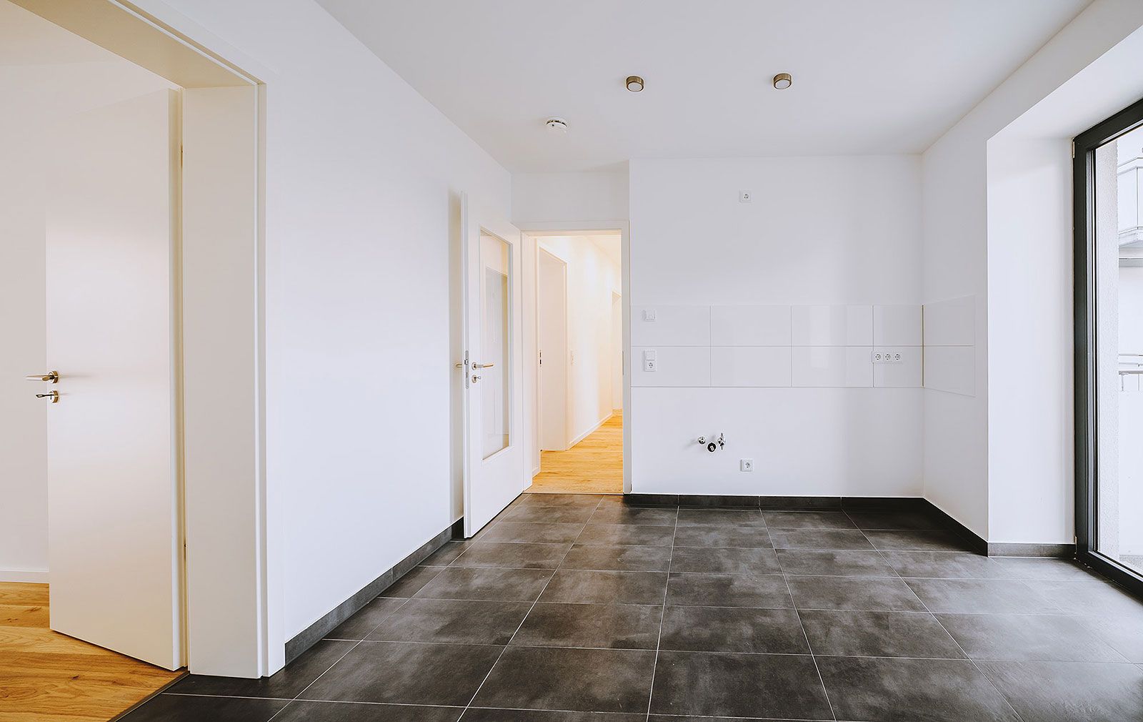 Leerer Küchenraum mit dunklem Fliesenboden, weißen Wänden und Anschlüssen für Küchenzeile in moderner Neubauwohnung