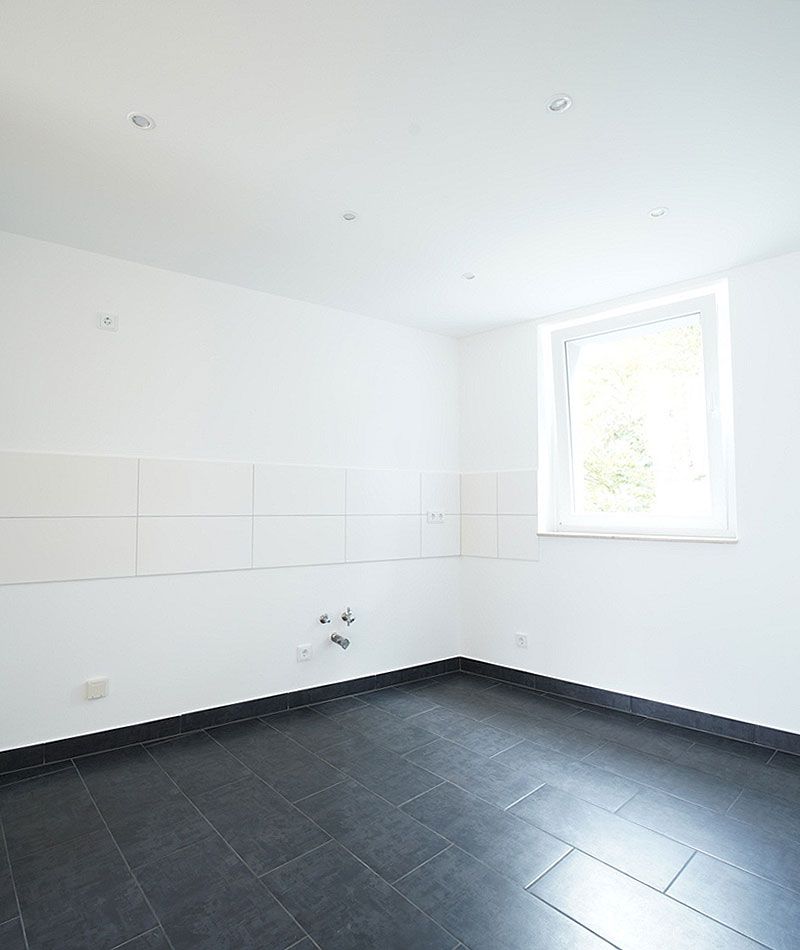Helle, unmöblierte Küche mit weißen Wänden, dunklem Fliesenboden und Anschlüssen für Spüle und Herd