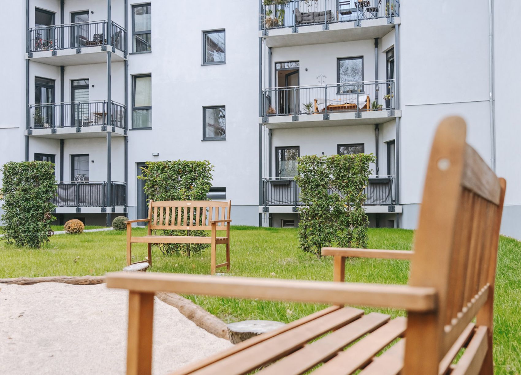 Grüner Innenhof mit Holzbänken, Hecken und Sandkasten vor einem modernen Mehrfamilienhaus mit Balkonen
