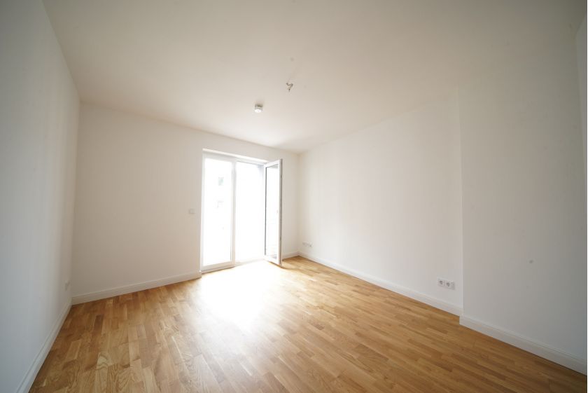 Helles, leeres Zimmer mit Eichenparkett, weißen Wänden und bodentiefem Fenster mit Zugang zum Balkon in Neubauwohnung