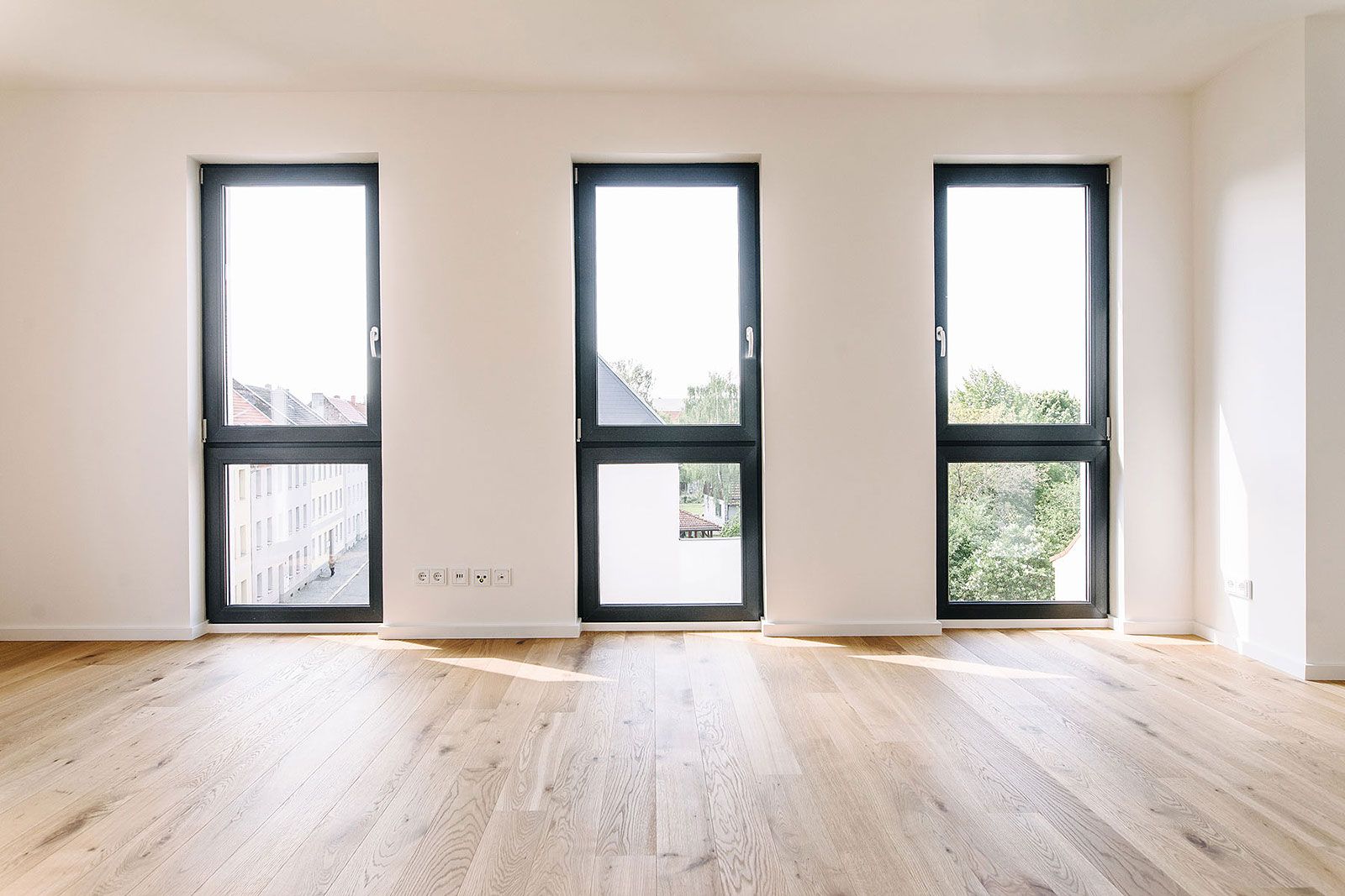 Helles Zimmer mit drei bodentiefen Fenstern, Parkettboden und Blick auf Stadt und Grünflächen
