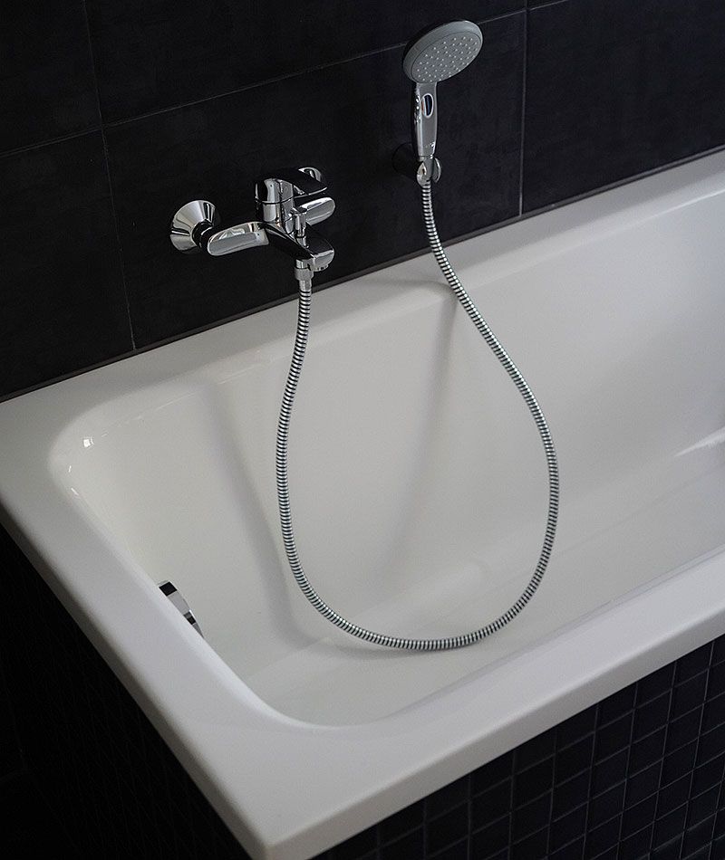 Weiße Badewanne mit Wandarmatur und Handbrause vor dunkler Fliesenwand in modernem Badezimmer