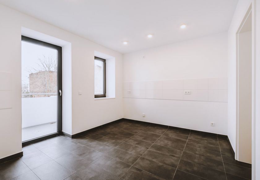 Leere Küche mit dunklem Fliesenboden, weißen Wänden, Fenster, Balkonzugang und Anschlüssen für Küchenzeile