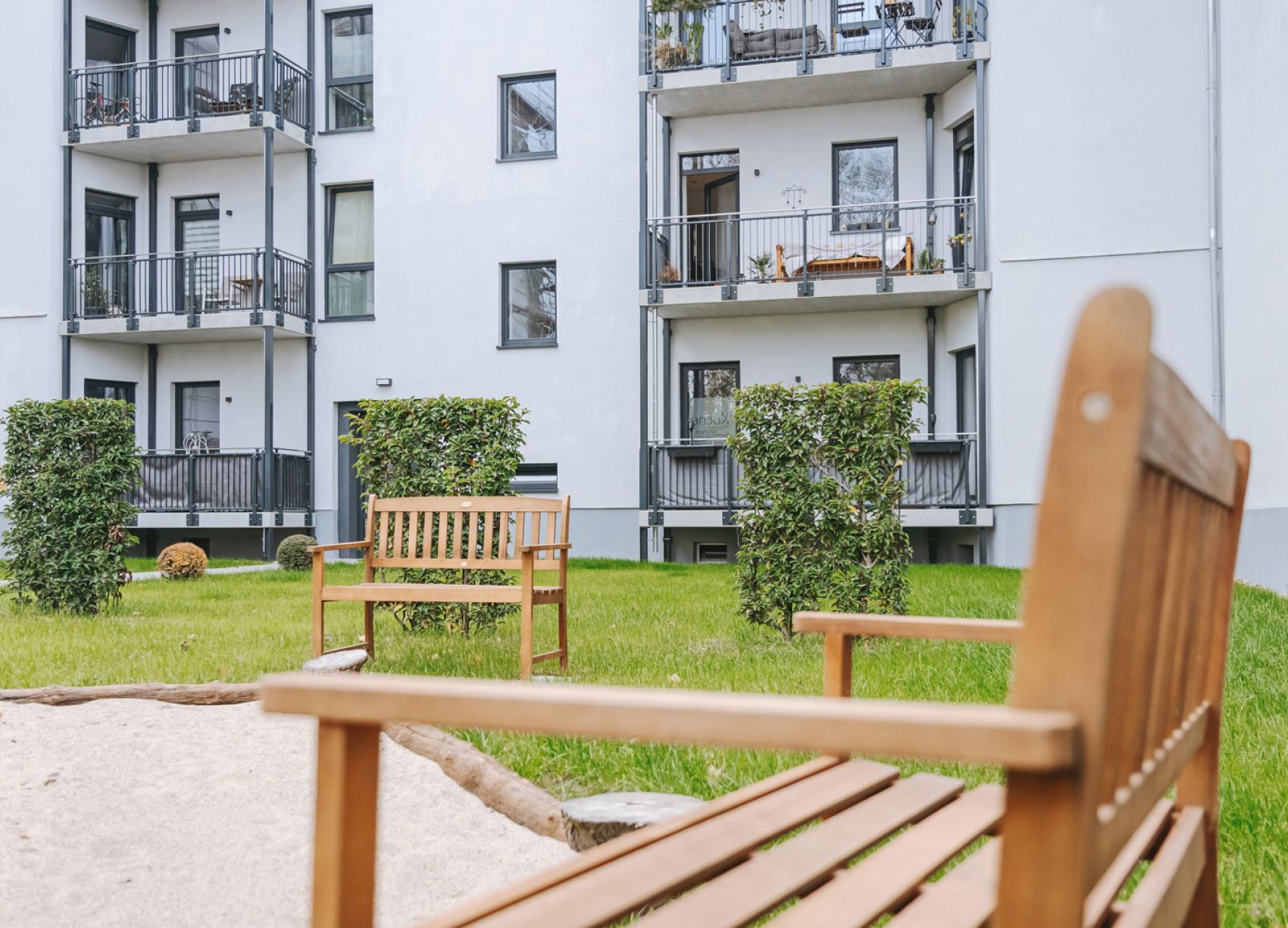 Grüner Innenhof mit Holzbänken, Hecken und Sandkasten vor einem modernen Mehrfamilienhaus mit Balkonen