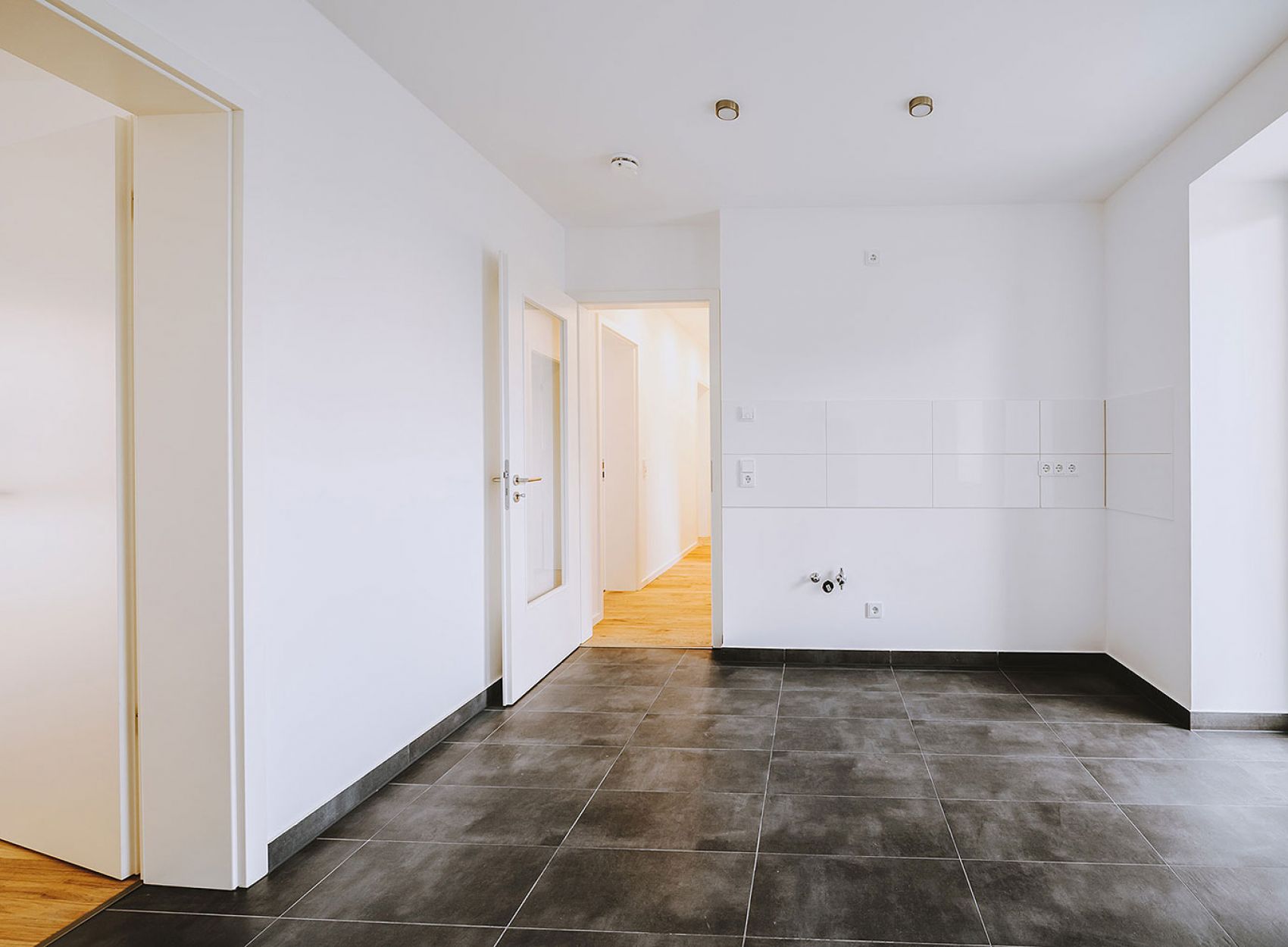 Leere, helle Küche mit dunklem Fliesenboden, weißen Wänden, Tür zum Balkon und Anschlüssen für Küchenmöbel