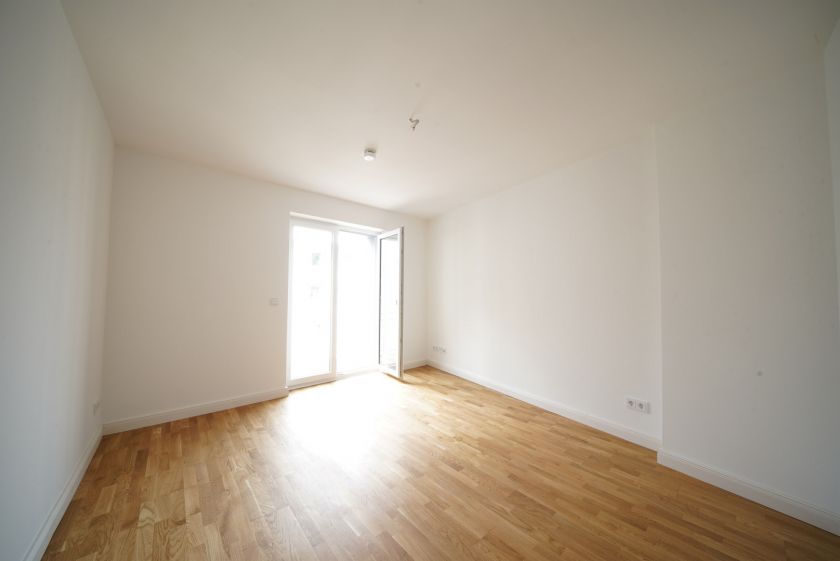 Helles, leeres Zimmer mit Eichenparkett, weißen Wänden und bodentiefem Fenster mit Zugang zum Balkon in Neubauwohnung